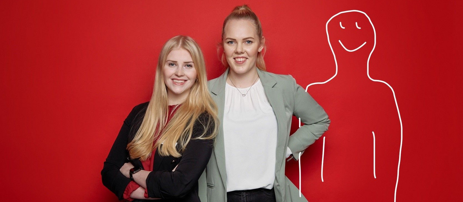 Pia Petersen und Lara Schomaker vor einer roten Wand. Rechts neben ihnen ist sein Männchen in weiß auf die Wand gemalt.