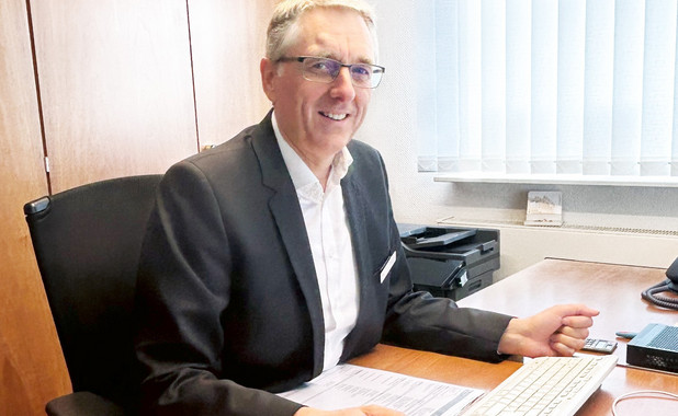 Stephan Röhr an seinem Schreibtisch im Büro. Er trägt einen Anzug und eine Brille und lächelt freundlich.