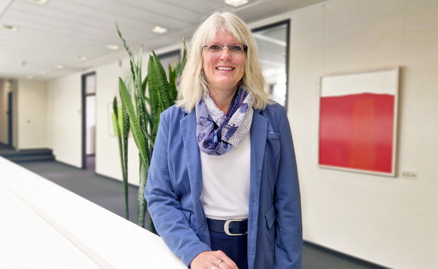 Carina Carstensen auf dem hell beleuchteten Flur einer Bankfiliale in einem blauen Blazer mit weißem Shirt. Sie trägt eine Brille und lächelt freundlich.