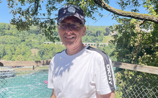 Stephan Röhr draußen im Grünen vor einem strahlend blauen Gewässer. Er trägt eine Schirmmütze, eine Brille und ein weißes T-Shirt.