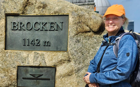 Carina Carstensen neben einem großen Felsen auf dem Brocken 1142 Meter steht. Sie trägt eine blaue Jacke, einen Rucksack und eine orangefarbene Schirmmütze.