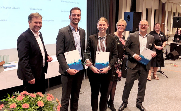 Sieben Personen auf der Abschlussfeier in einer großen Halle vor einer an eine Wand projizierte Präsentation. Sie tragen Abendkleidung und einige halten ein Exemplar eines bestimmten Buches in den Händen.