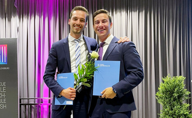 Asmus Schmidt und ein anderer junger Mann in Anzügen auf einer Bühne mit ihren Abschlusszeugnismappen in der Hand. Sie freuen sich sichtlich und lächeln in die Kamera.