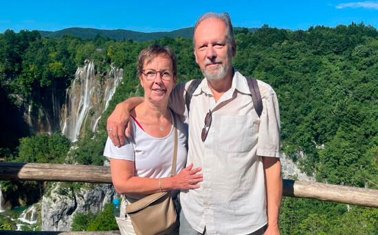 Waldemar Petarus und seine Frau vor einem großen Wasserfall. Sie tragen T-Shirts, die Sonne scheint und der Himmel ist strahlend blau.