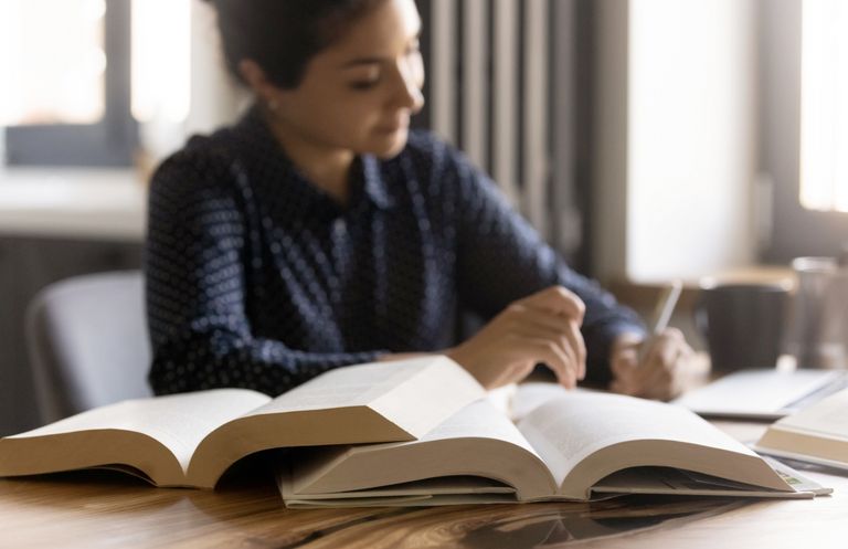Eine Frau sitzt an einem Schreibtisch und schreibt etwas auf. Neben ihr liegen mehrere aufgeschlagene Bücher.
